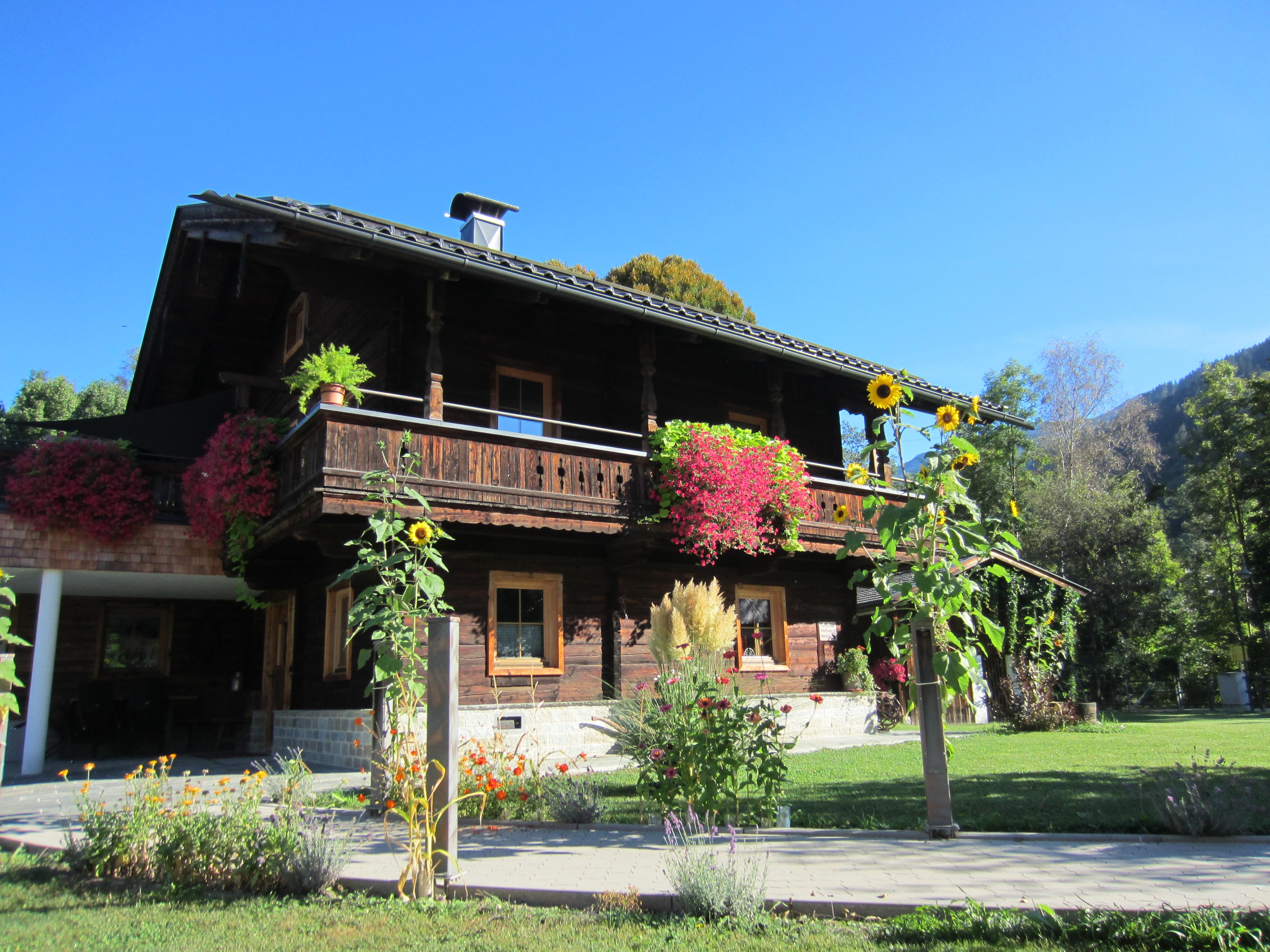 Ein Holzhaus mit Balkon, umgeben von bunten Blumen und Sonnenblumen, vor einem blauen Himmel und grünen Bäumen.