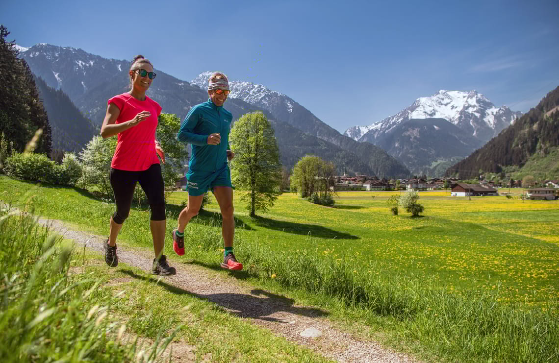 Easy Trail Mayrhofen ©Dominic Ebenbichler Two runners on the Easy Trail valley path in Mayrhofen-Hippach, surrounded by blooming meadows, alpine landscape, and snow-covered mountains in bright sunshine.