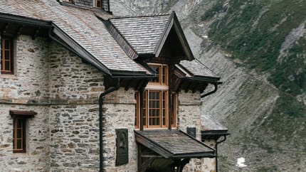 Stone mountain house with wooden windows, surrounded by snow-capped mountains and greenery in the valley.