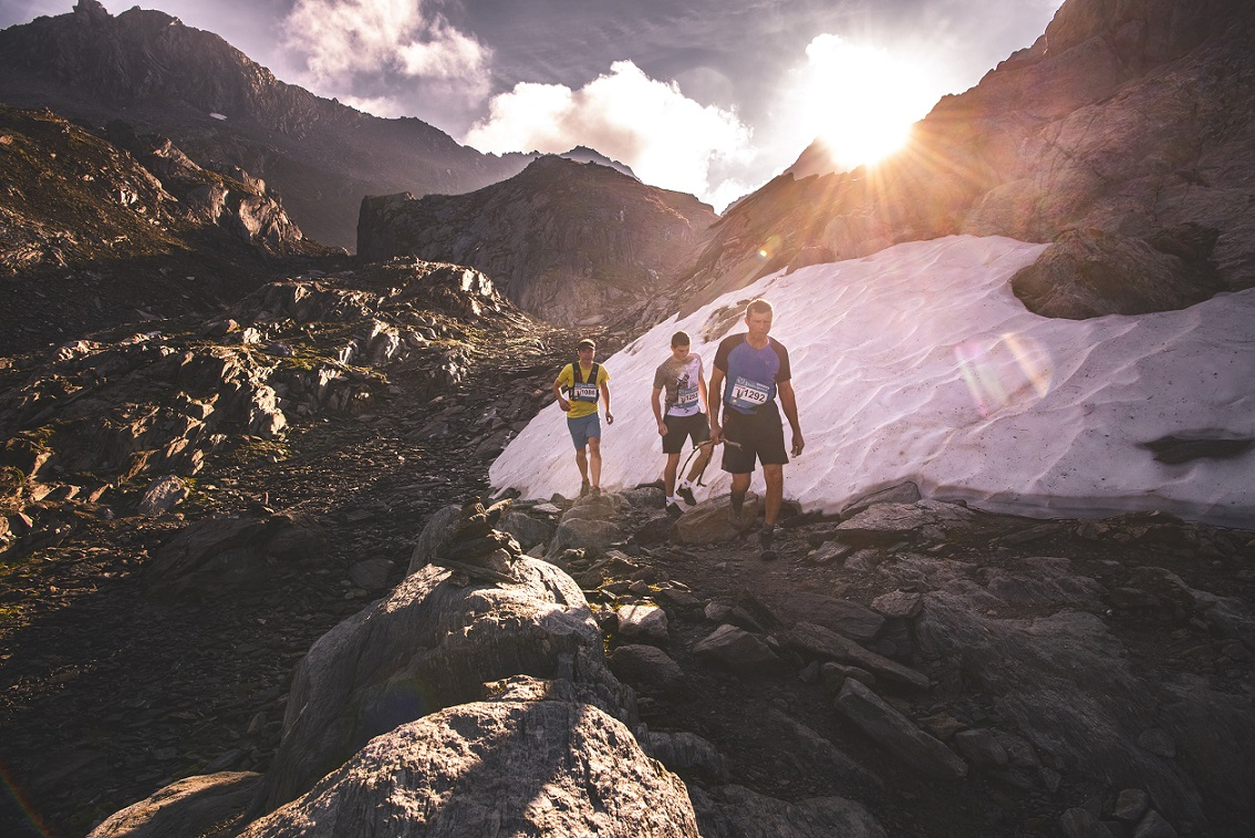 The picture shows three runners in alpine terrain with a snowfield in the background.