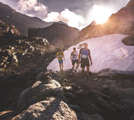 The picture shows three runners in alpine terrain with a snowfield in the background.