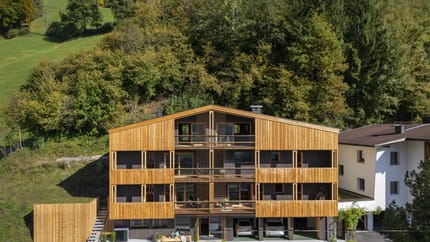 Modern wooden house with balcony, set against green trees and mountains, with parking below.