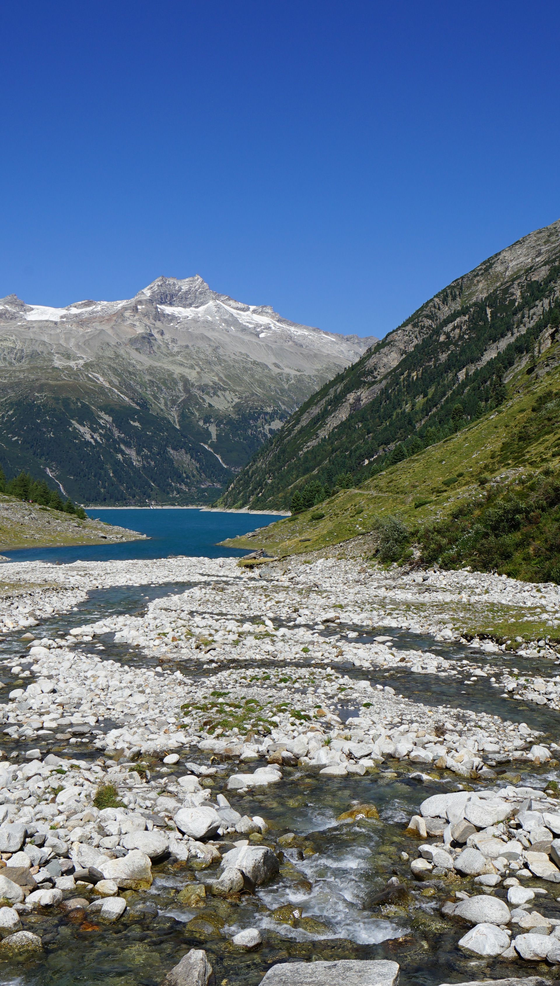 Zillertal Alps Nature Park Schlegeis reservoir in the Zillertal Alps Nature Park, surrounded by green alpine meadows and high mountains with remnants of snow. A mountain stream flows over gravel into the lake. An idyllic destination in the Mayrhofen-Hippach holiday region.