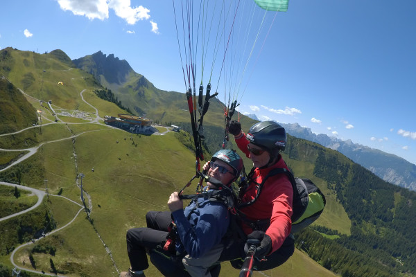Zwei Paraglider fliegen über grüne Hügel und Berge bei sonnigem Himmel.