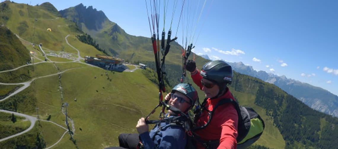 Two paragliders flying over green hills and mountains on a sunny day.