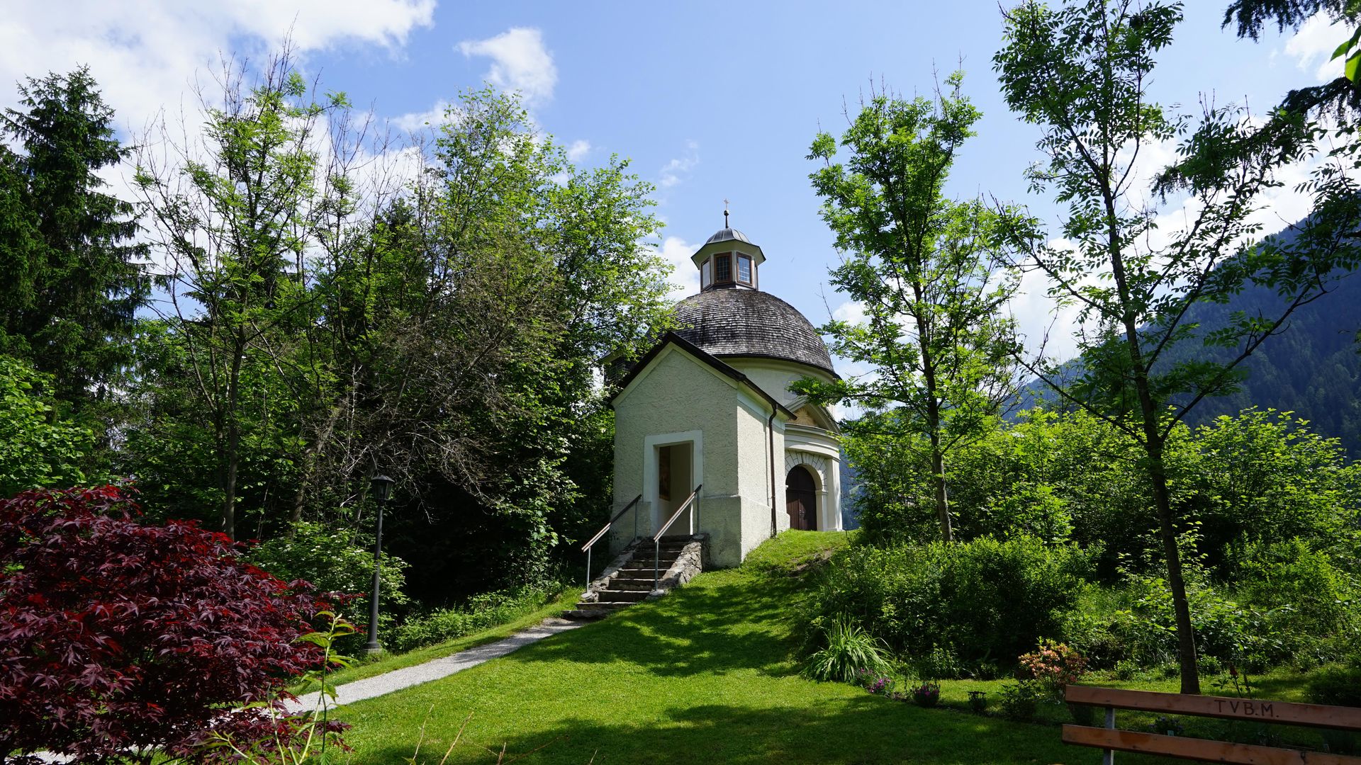 Die Kapelle Burgschrofen steht inmitten einer grünen Gartenlandschaft, umgeben von Bäumen, Sträuchern und Blumen. Im Hintergrund erheben sich Berge unter blauem Himmel.