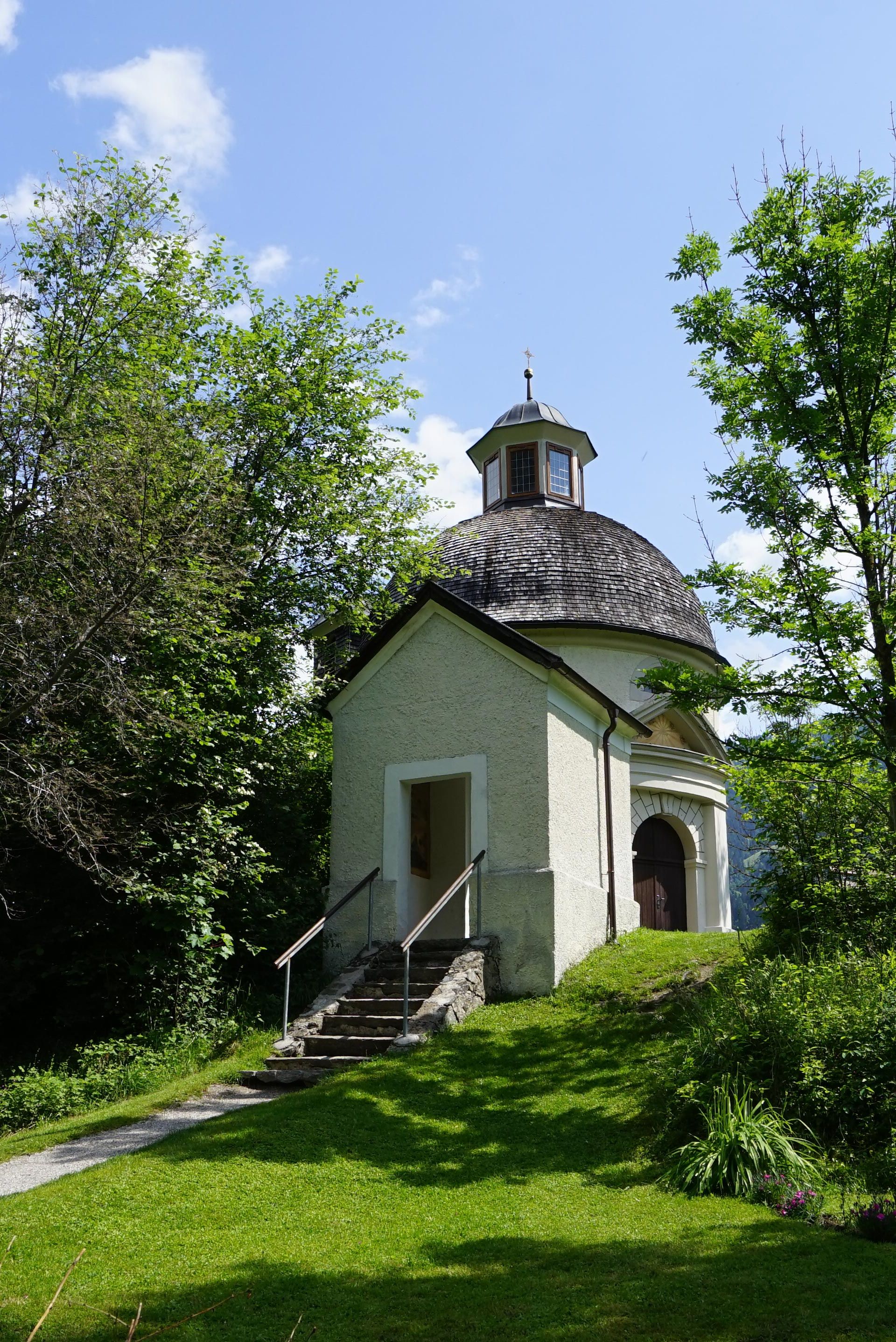 Die Kapelle Burgschrofen steht inmitten einer grünen Gartenlandschaft, umgeben von Bäumen, Sträuchern und Blumen. Im Hintergrund erheben sich Berge unter blauem Himmel.