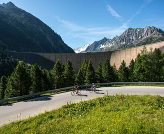 Biketour zum Schlegeisspeicher im Zillertal: Zwei Radfahrer fahren auf der kurvigen Schlegeis Alpenstraße durch grüne Berglandschaft. Im Hintergrund erhebt sich die hohe Staumauer zwischen bewaldeten Hängen und schroffen Gipfeln in den blauen Himmel.