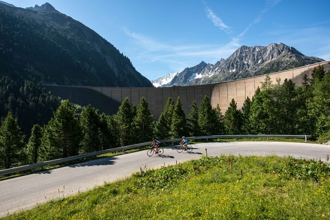Biketour Schlegeis ©Michael Werlberger Bike tour to the Schlegeis Reservoir in Zillertal: Two cyclists ride along the winding Schlegeis Alpine Road through green mountain scenery. In the background, the tall dam rises between forested slopes and rugged peaks into the blue sky.