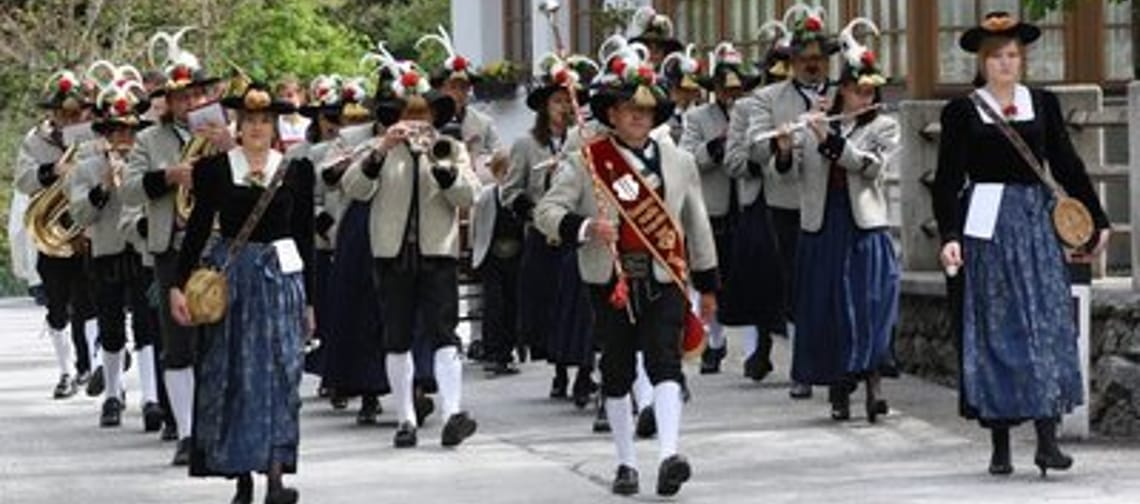 The image shows a brass band in traditional attire marching in a festive parade through a street. The musicians wear decorated hats and play various brass instruments.