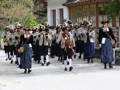 Das Bild zeigt eine Blaskapelle in traditioneller Tracht, die in einem festlichen Umzug durch eine Straße marschiert. Die Musiker tragen geschmückte Hüte und spielen verschiedene Blasinstrumente.