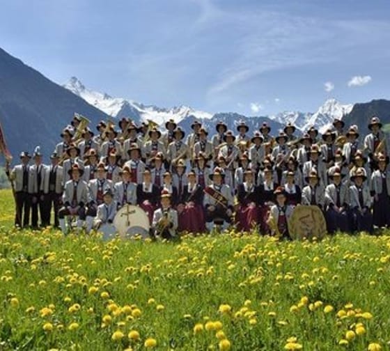 The brass band poses in traditional attire on a blooming meadow with an impressive mountain backdrop for a group photo.