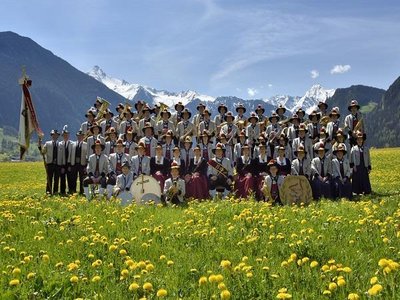 The brass band poses in traditional attire on a blooming meadow with an impressive mountain backdrop for a group photo.