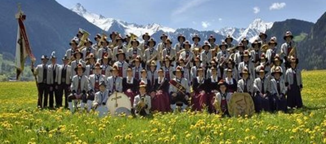 Die Blaskapelle posiert in traditioneller Tracht auf einer blühenden Wiese vor einer beeindruckenden Bergkulisse für ein Gruppenfoto.