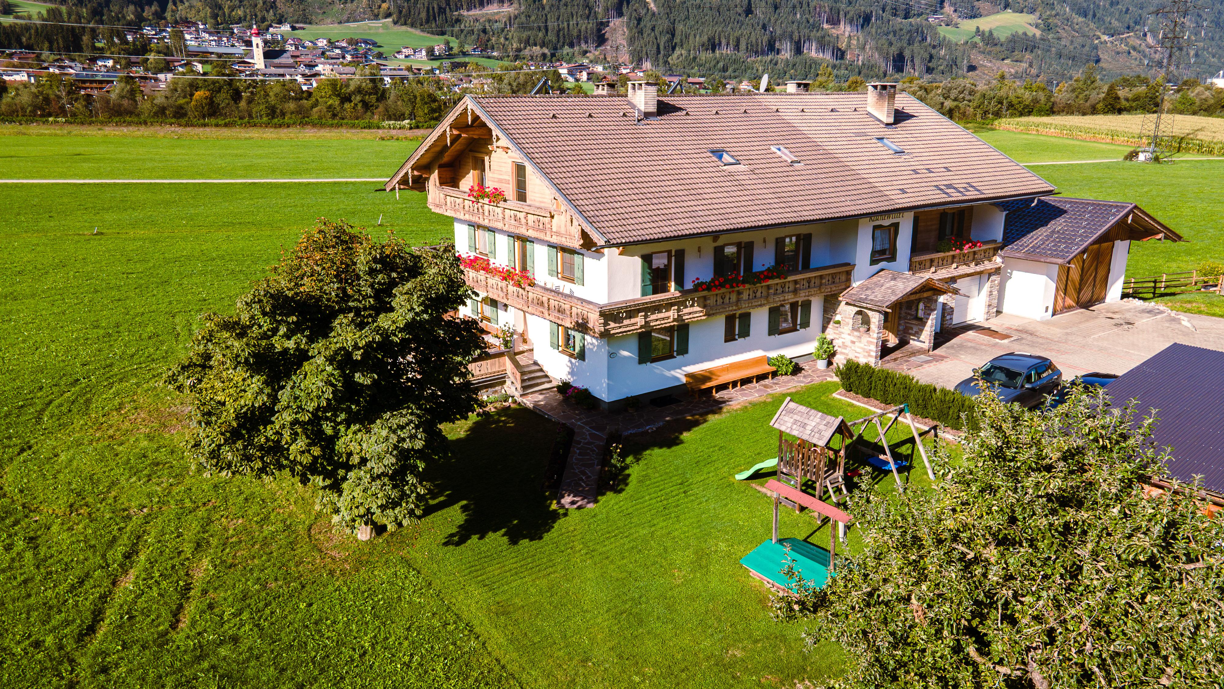 House on a green field with playground and trees, with a small town and hills in the background.