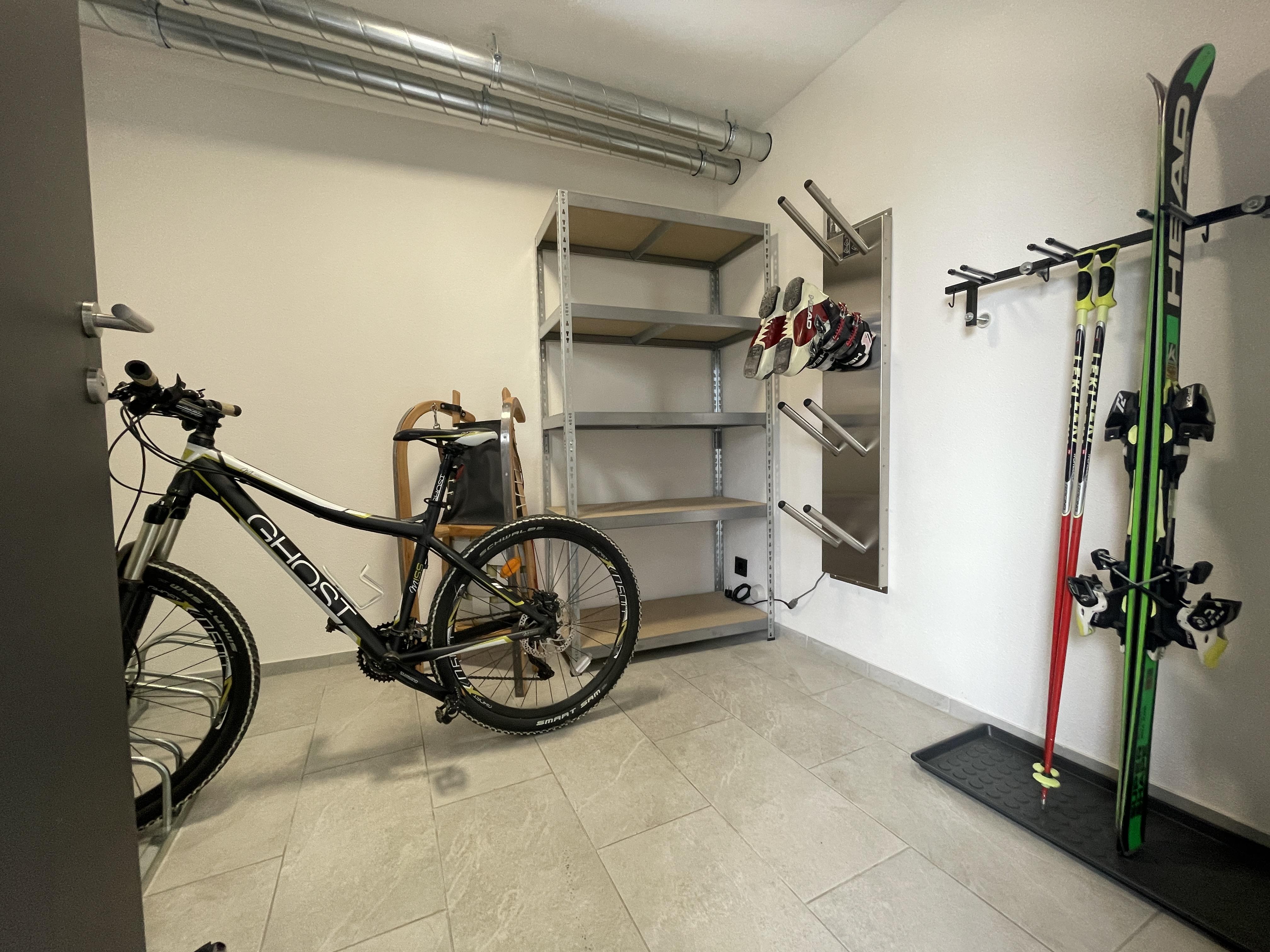 Fahrrad, Skier und Schuhe in einem Abstellraum mit Regal und Wandhaken, weiße Wände und Fliesenboden.