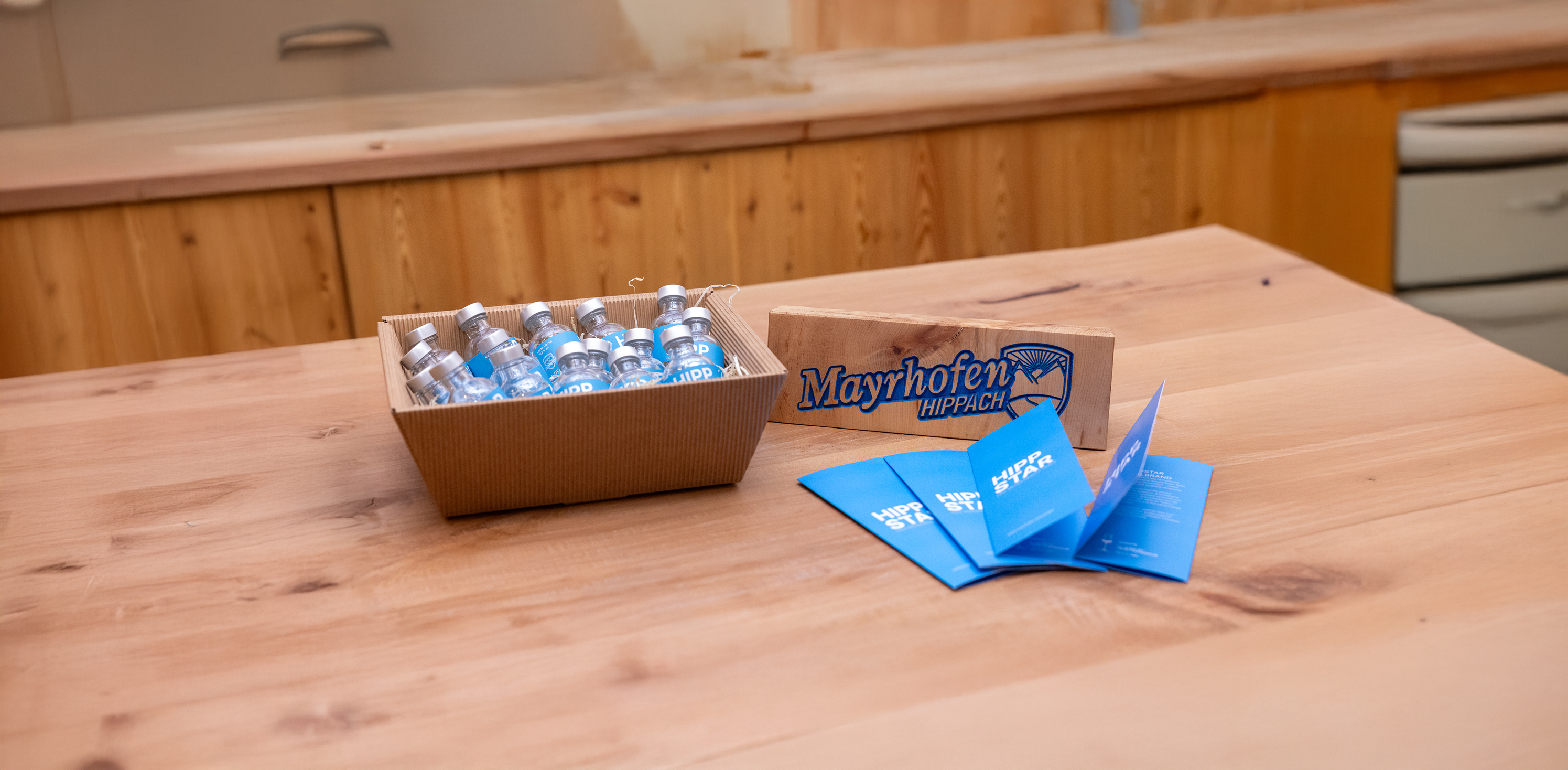 Karton mit kleinen Glasflaschen, silbernen Verschlüssen und blauen Etiketten mit weißer Schrift „HIPP", daneben ein Holzschild mit der Aufschrift „Mayrhofen-Hippach" und Logo sowie gefächerte blaue Flyer mit weißer Schrift „HIPPSTAR" auf einem Holztisch.