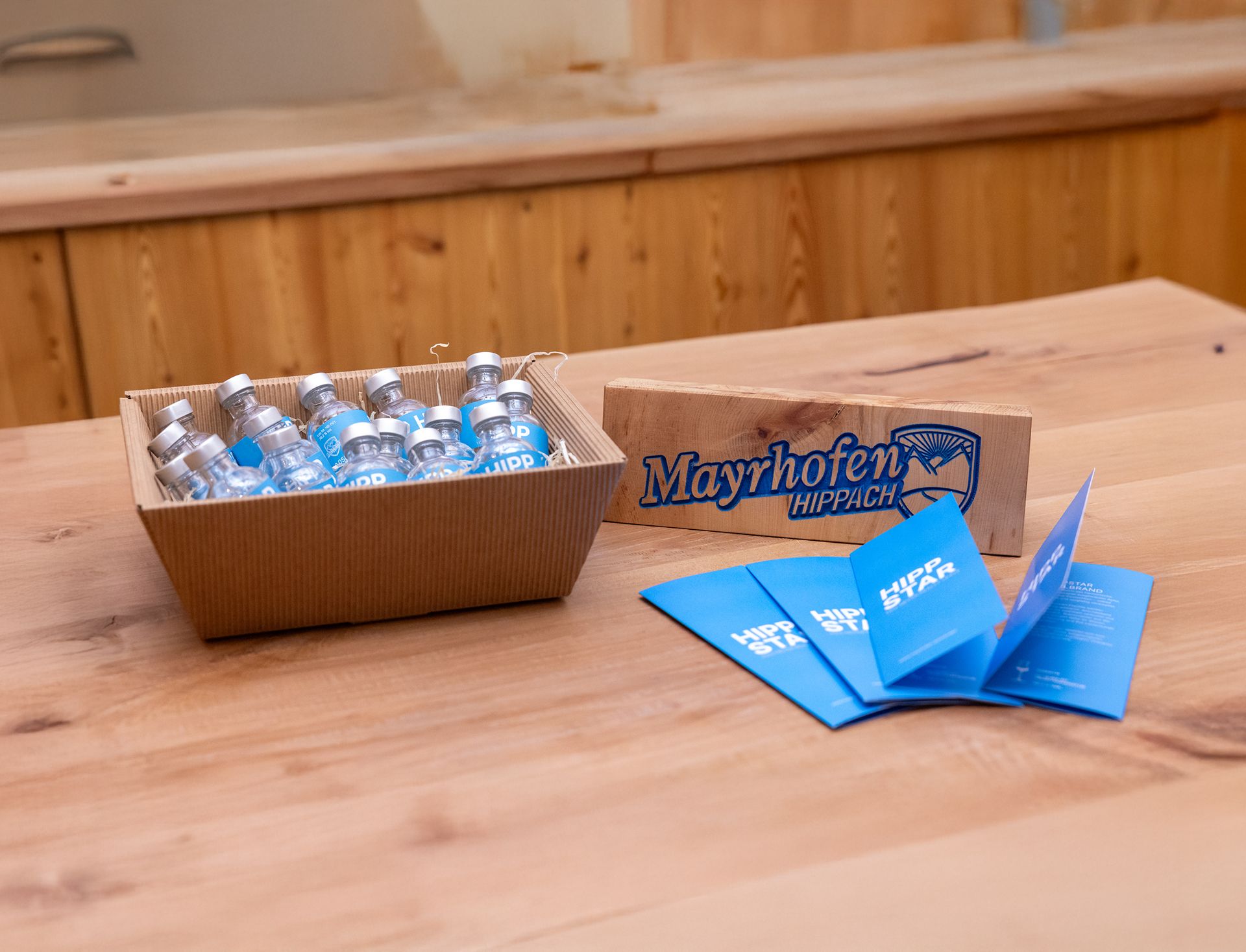 Karton mit kleinen Glasflaschen, silbernen Verschlüssen und blauen Etiketten mit weißer Schrift „HIPP", daneben ein Holzschild mit der Aufschrift „Mayrhofen-Hippach" und Logo sowie gefächerte blaue Flyer mit weißer Schrift „HIPPSTAR" auf einem Holztisch.