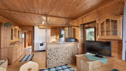 Innenansicht eines Holzhauses mit Küche, Holzdecken, Möbel und Fernseher, gemütlich und rustikal gestaltet.