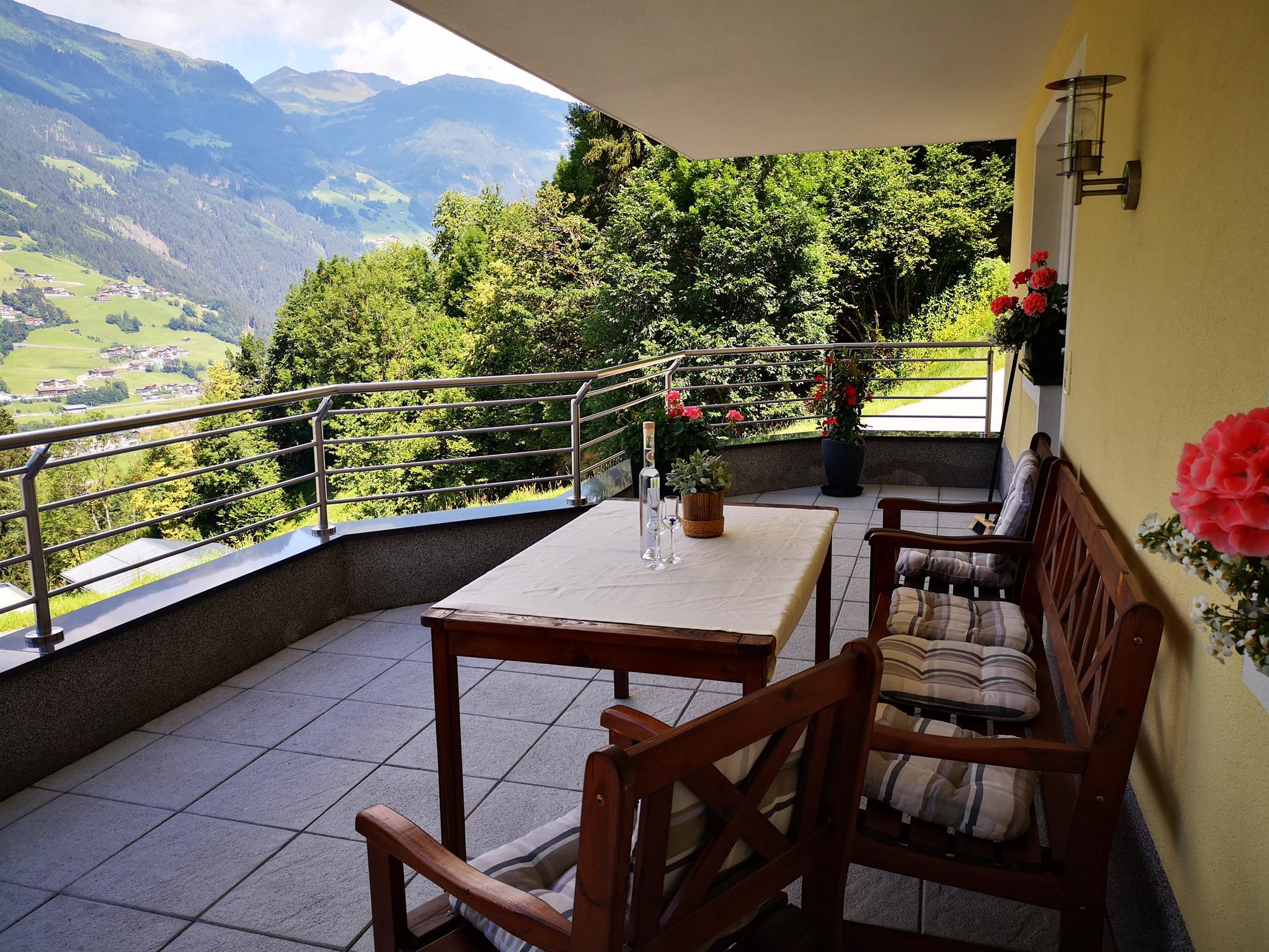 Balcony with wooden furniture, flowers, and a view of green hills and mountains.