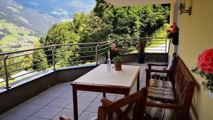 Balcony with wooden furniture, flowers, and a view of green hills and mountains.