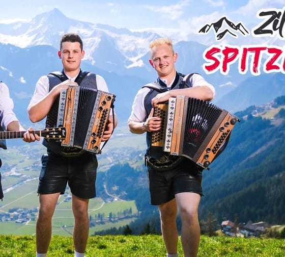 Drei junge Männer mit traditionellen Trachten, spielen Gitarre und Akkordeon vor Bergkulisse, Titel: Zillertaler Spitzbuam