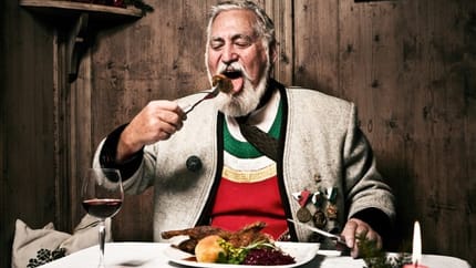 The picture shows a man wearing traditional Zillertal costume, dining in a cozy, old-fashioned room. In front of him is a glass of red wine, and a red candle provides warm, atmospheric lighting.