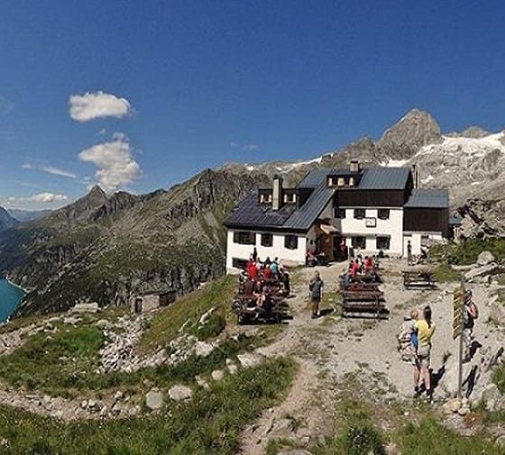 Mountain hut with people, surrounded by mountains and a lake in the background, on a sunny day.