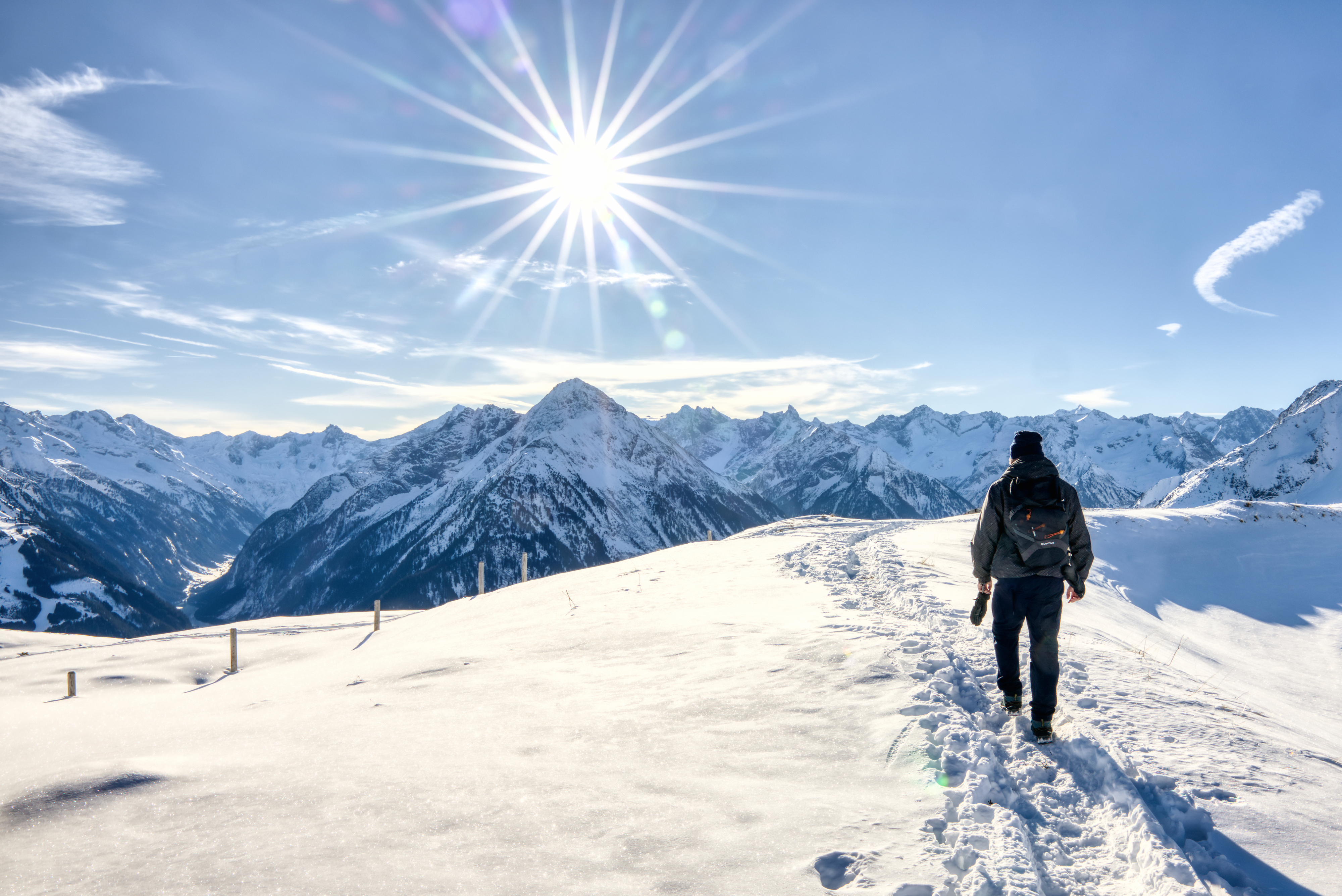 In einer verschneiten Bergwelt ist eine Dame bei strahlendem Sonnenschein unterwegs. Die Dame sieht man von hinten, wie sie den bereits vorhandenen Spuren im Schnee folgt.Spuren im Schnee  von hinten sieht 