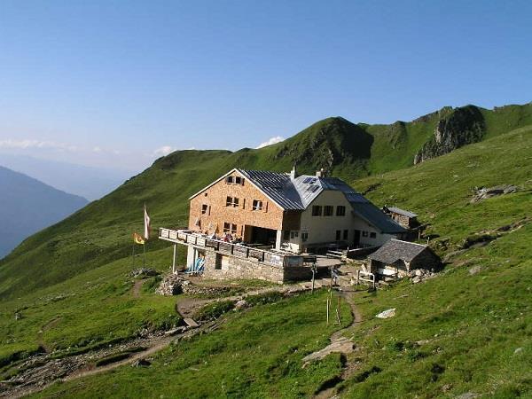 Hütten auf grünem Berghang mit Weg, umgeben von Bergen bei klarem Himmel.