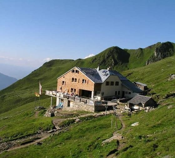 Hütten auf grünem Berghang mit Weg, umgeben von Bergen bei klarem Himmel.