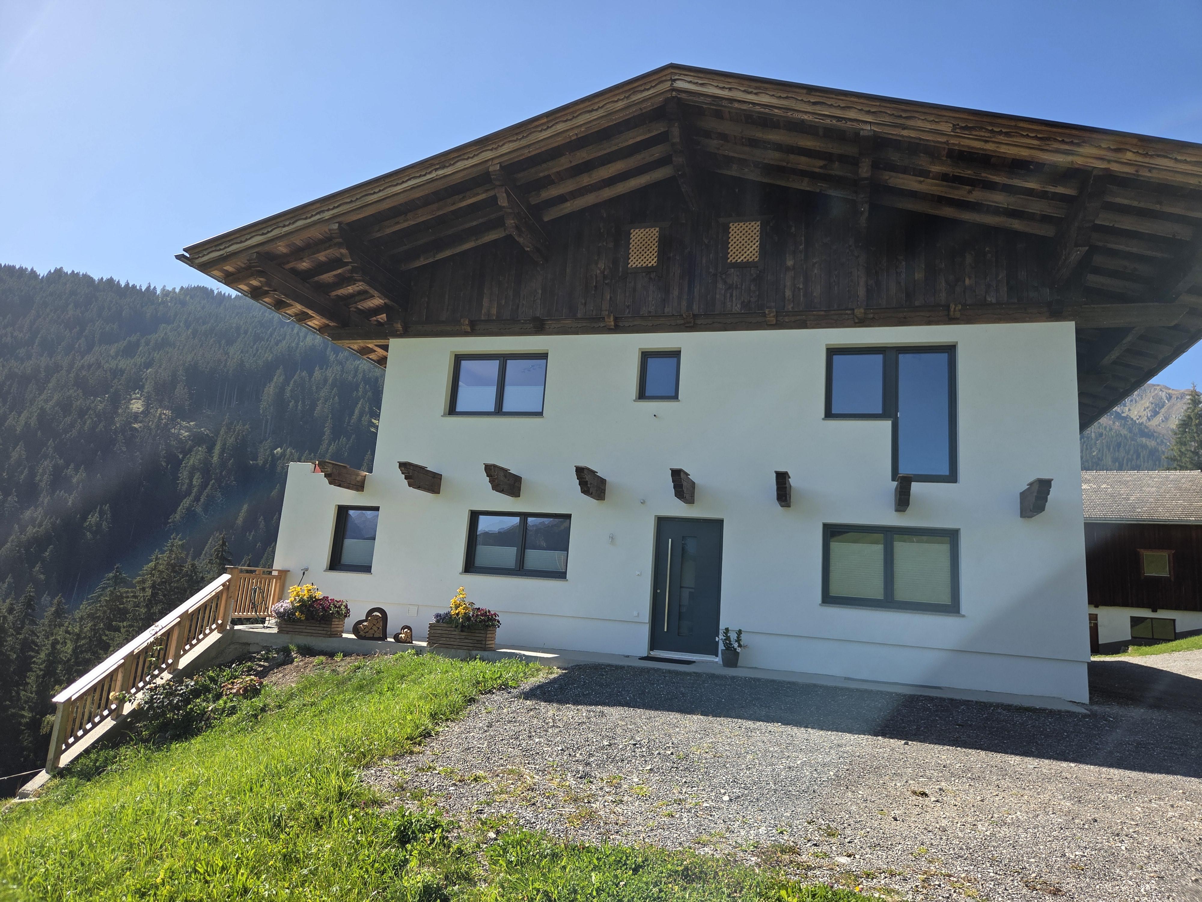 Modernes Haus mit weißer Fassade, dunklem Holzdach und Bergblick im Hintergrund, Blumen im Vordergrund.