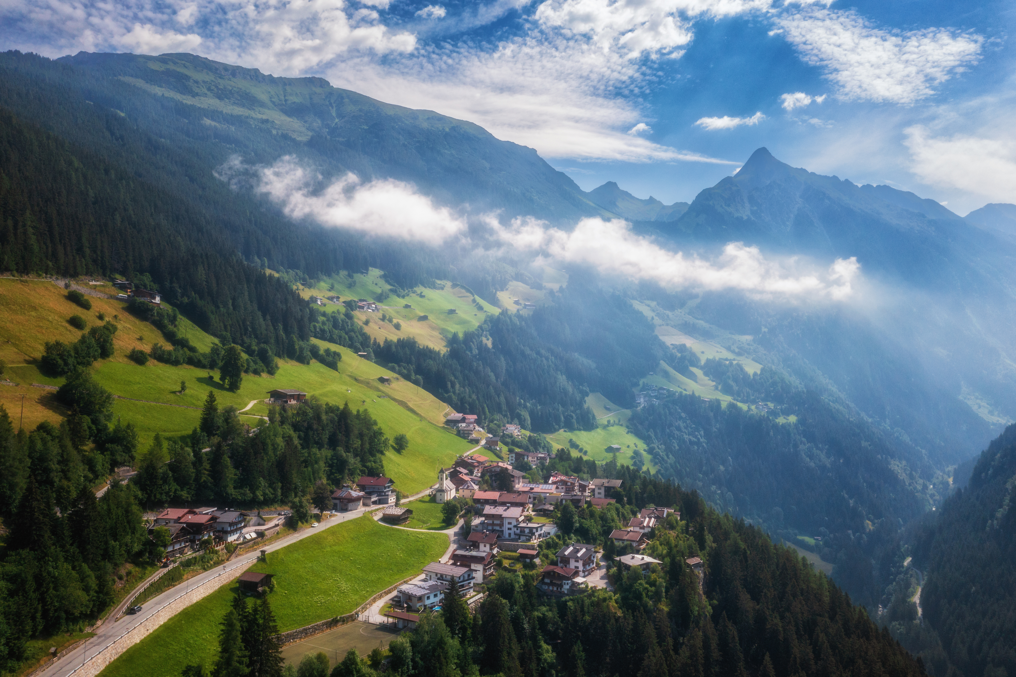 Summer aerial view of Brandberg village in Zillertal: Traditional alpine hamlet nestled on lush green meadows between forested mountain slopes. Morning mist drifts through the valley under dramatic cloudy skies with sun breaking through.