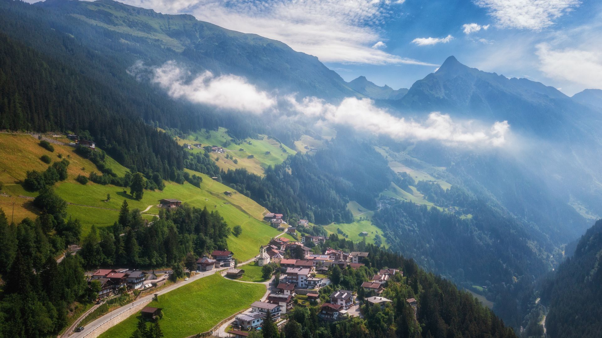 Summer aerial view of Brandberg village in Zillertal: Traditional alpine hamlet nestled on lush green meadows between forested mountain slopes. Morning mist drifts through the valley under dramatic cloudy skies with sun breaking through.