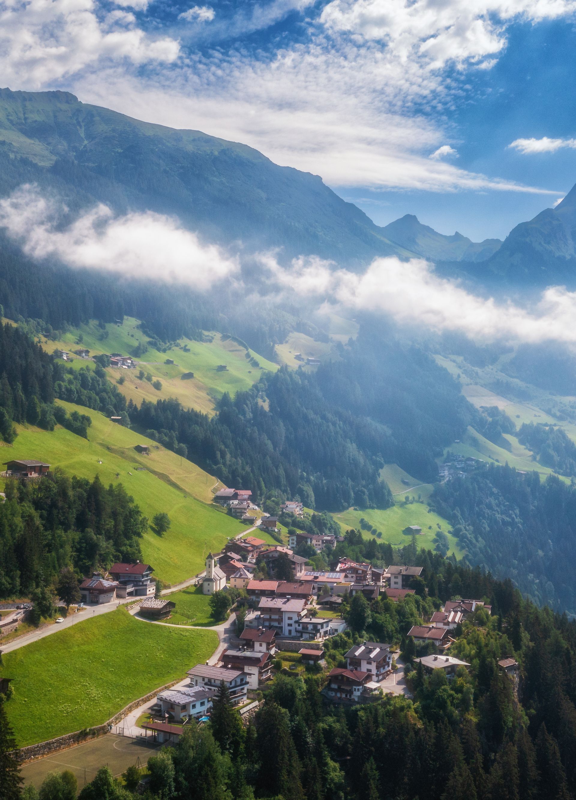 Summer aerial view of Brandberg village in Zillertal: Traditional alpine hamlet nestled on lush green meadows between forested mountain slopes. Morning mist drifts through the valley under dramatic cloudy skies with sun breaking through.