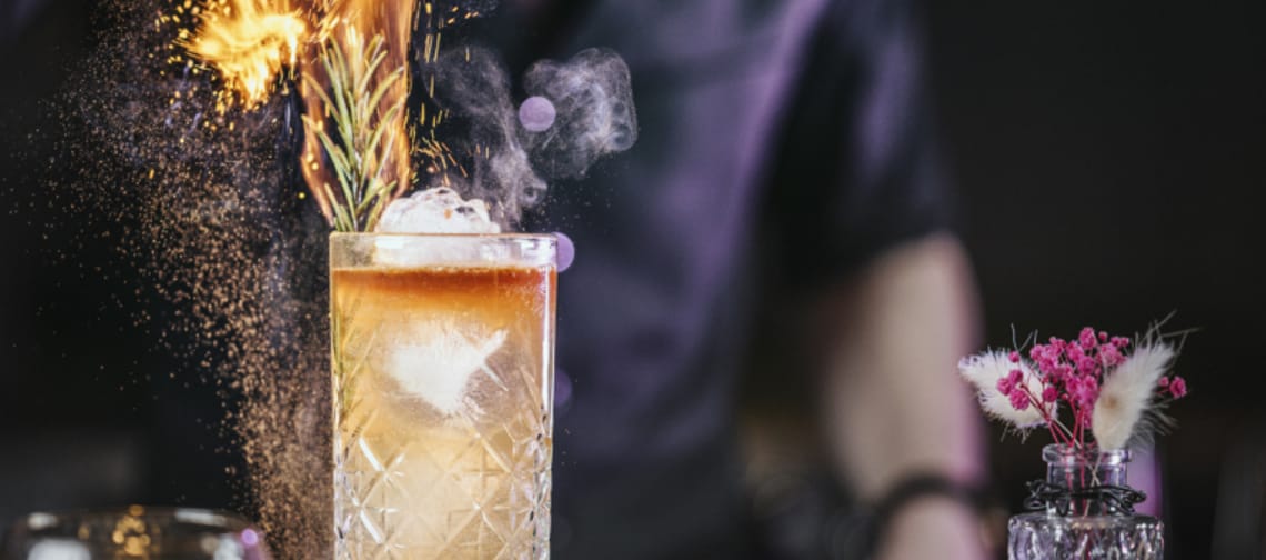 Das Bild zeigt einen Cocktail, aus dem Flammen aufsteigen – ein echter Blickfang. Der Hintergrund ist schwarz, wodurch die lodernden Flammen besonders eindrucksvoll zur Geltung kommen