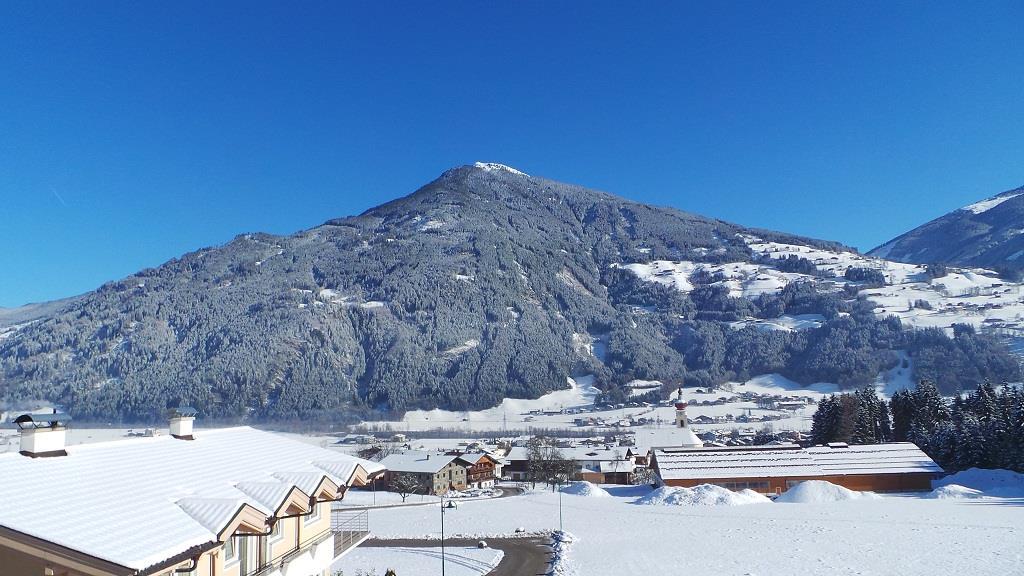 Snow-covered houses with a large forested mountain in the background under a clear blue sky.