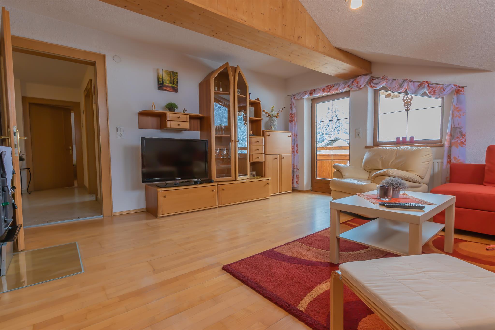 Wohnzimmer mit Holzmöbeln, Sofa, Fernseher, Fenster mit Blumenmuster-Vorhängen, Holzbalken an der Decke.