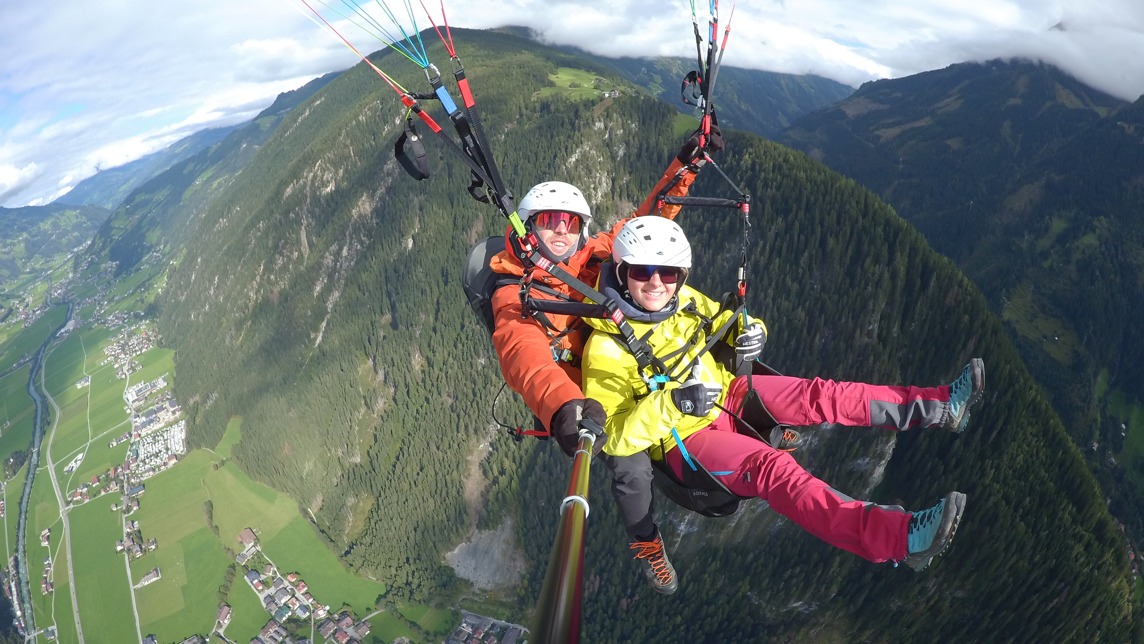 The image shows a paraglider high up in the air during a tandem flight. Mountains and green meadows in the background.