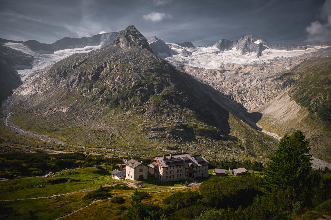 Berliner Hütte im Zillertal – Berghütte am Fuße der Zillertaler Alpen | Mayrhofen-Hippach Die Berliner Hütte im Zillertal: rustikale Steinbau-Hütte mit geschindeltem Dach in grüner Almwiese, dahinter schroffe Felsflanken und schneebedeckte Gipfel mit Gletscher unter dramatischen Wolken.