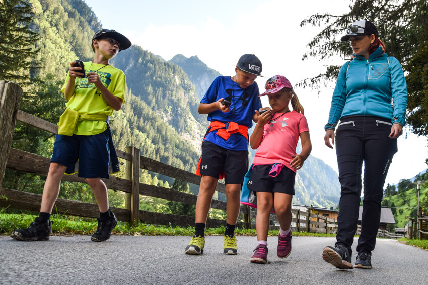 Geocache "Sagenweg" in Ginzling Auf Schatzsuche im Bergsteigerdorf im Zillertal – Geocache Sagenweg in Ginzling, Tirol: Zwei Kinder mit GPS-Geräten und zwei Erwachsene gehen auf einem befestigten Weg. Neben ihnen ein Holzzaun, im Hintergrund grüne Wiesen, hohe Berge und dichter Wald.