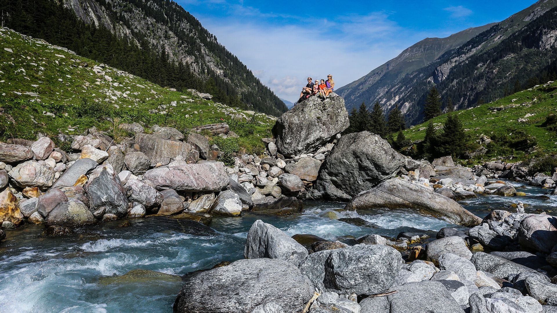 Family trip to the Stilluptal Valley Family sitting on a large rock by a mountain stream in the Stilluptal near Mayrhofen. Surrounded by green meadows, boulders, and steep mountain slopes. The stream rushes through the alpine valley under a blue sky.