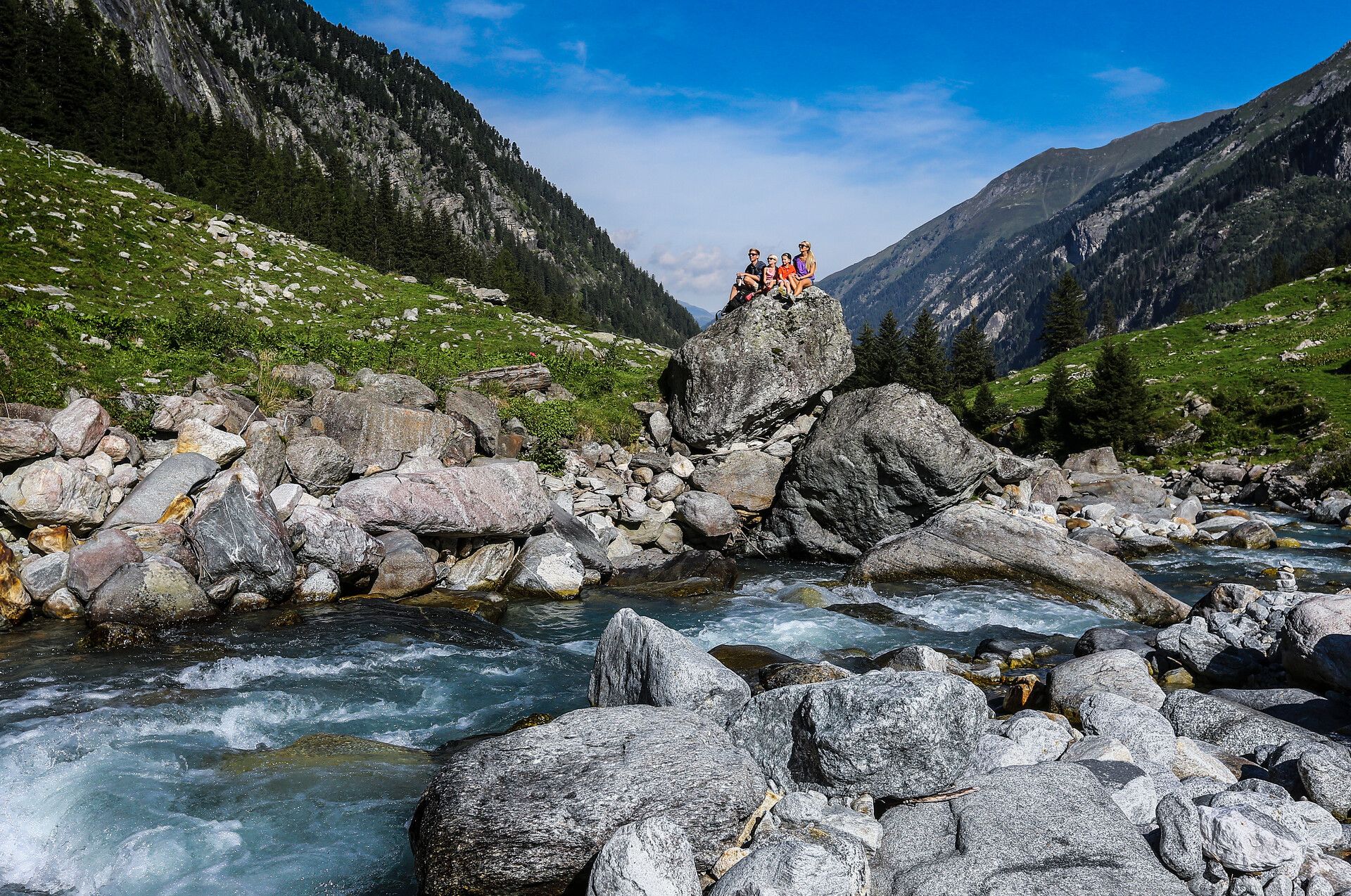Family trip to the Stilluptal Valley Family sitting on a large rock by a mountain stream in the Stilluptal near Mayrhofen. Surrounded by green meadows, boulders, and steep mountain slopes. The stream rushes through the alpine valley under a blue sky.