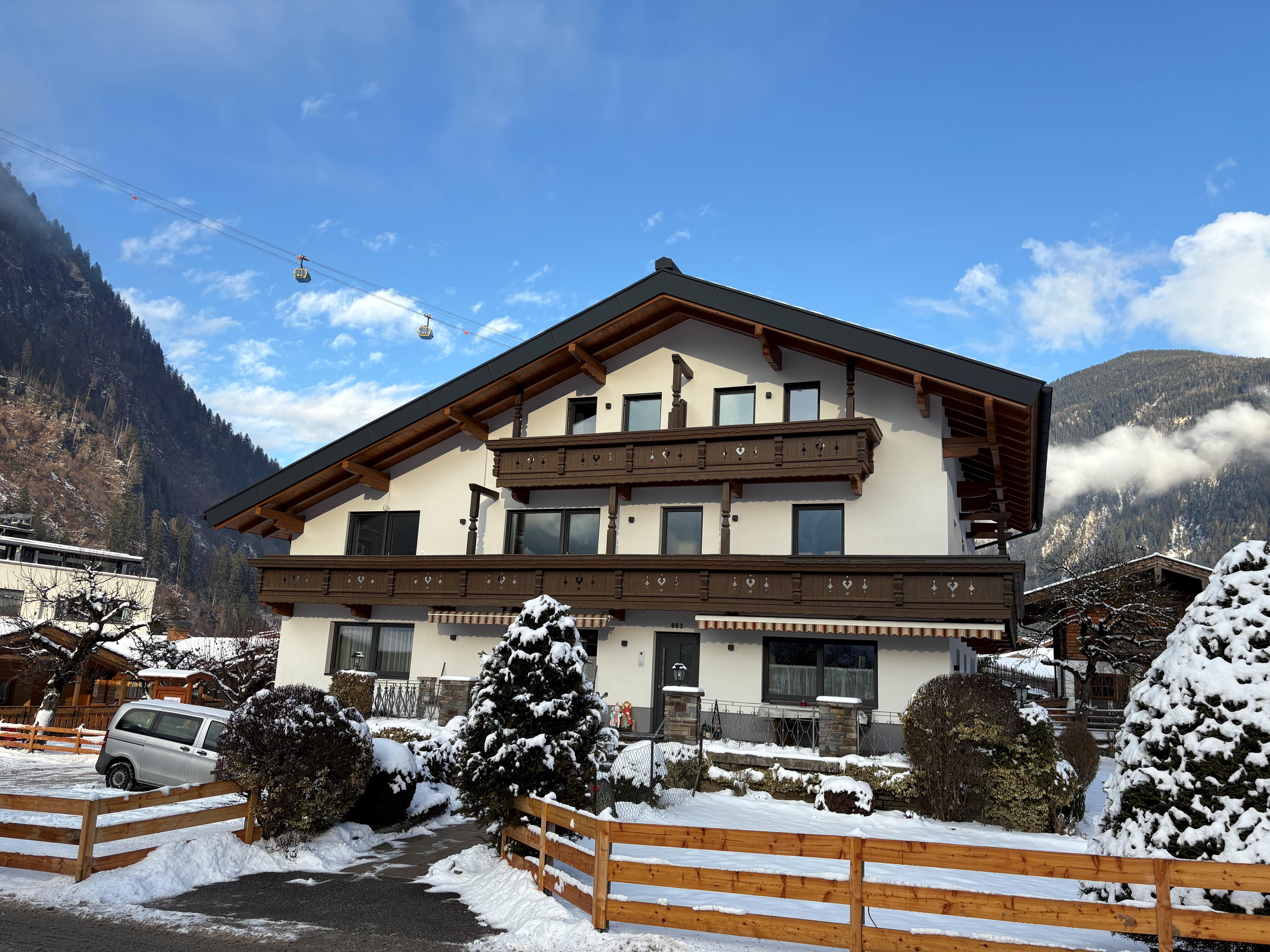 Ein modernes Haus mit Holzdetails in einer verschneiten Berglandschaft, darunter eine Gondelbahn am Himmel.