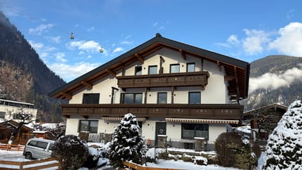 Ein modernes Haus mit Holzdetails in einer verschneiten Berglandschaft, darunter eine Gondelbahn am Himmel.