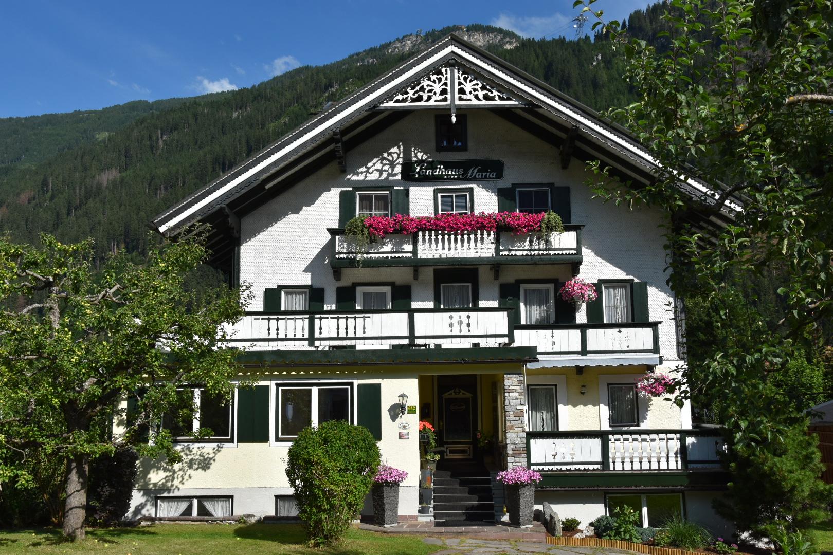 Alpine-style house with flower-filled balconies, surrounded by trees and mountains under a sunny sky.