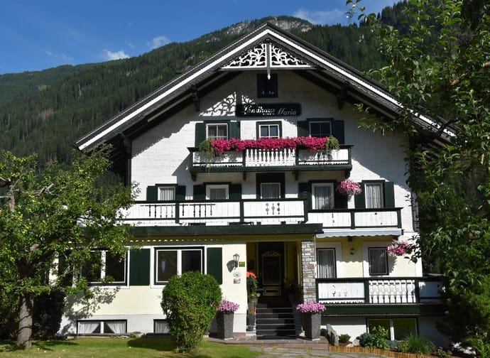 Alpine-style house with flower-filled balconies, surrounded by trees and mountains under a sunny sky.