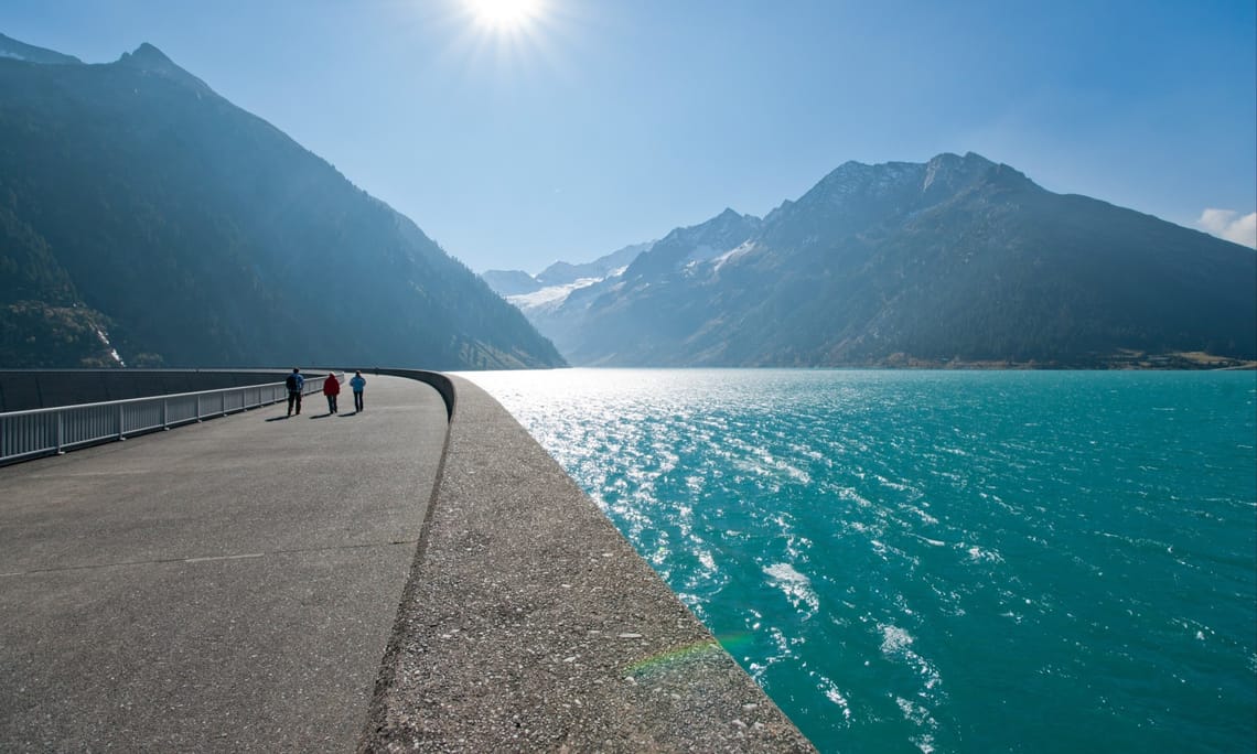 Schlegeis reservoir ©VERBUND Dam walkway at Schlegeis Reservoir in Zillertal, Tyrol. On the right, a turquoise lake; on the left, tall mountains. People walk in the sun. Snow-covered Alpine peaks rise in the background under a clear blue sky.