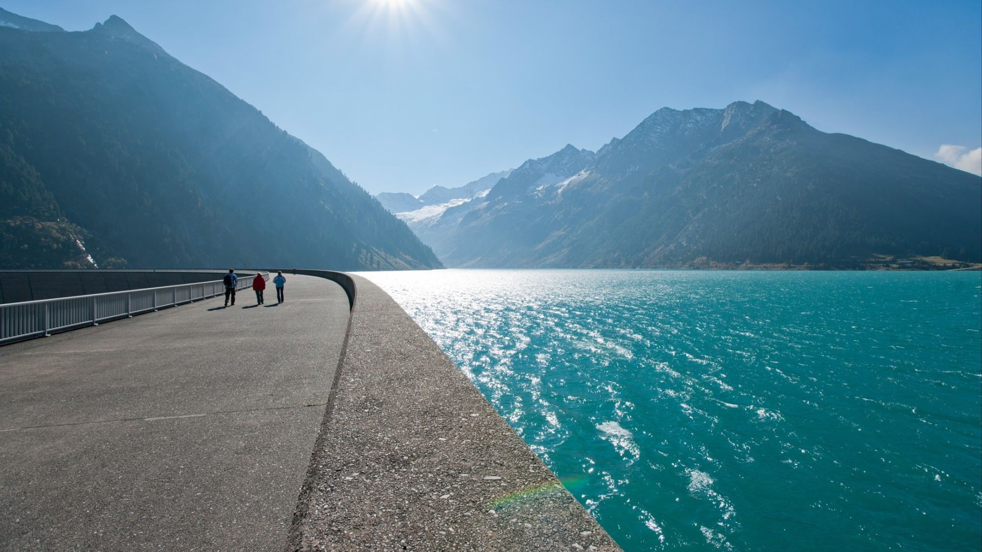Dam walkway at Schlegeis Reservoir in Zillertal, Tyrol. On the right, a turquoise lake; on the left, tall mountains. People walk in the sun. Snow-covered Alpine peaks rise in the background under a clear blue sky.