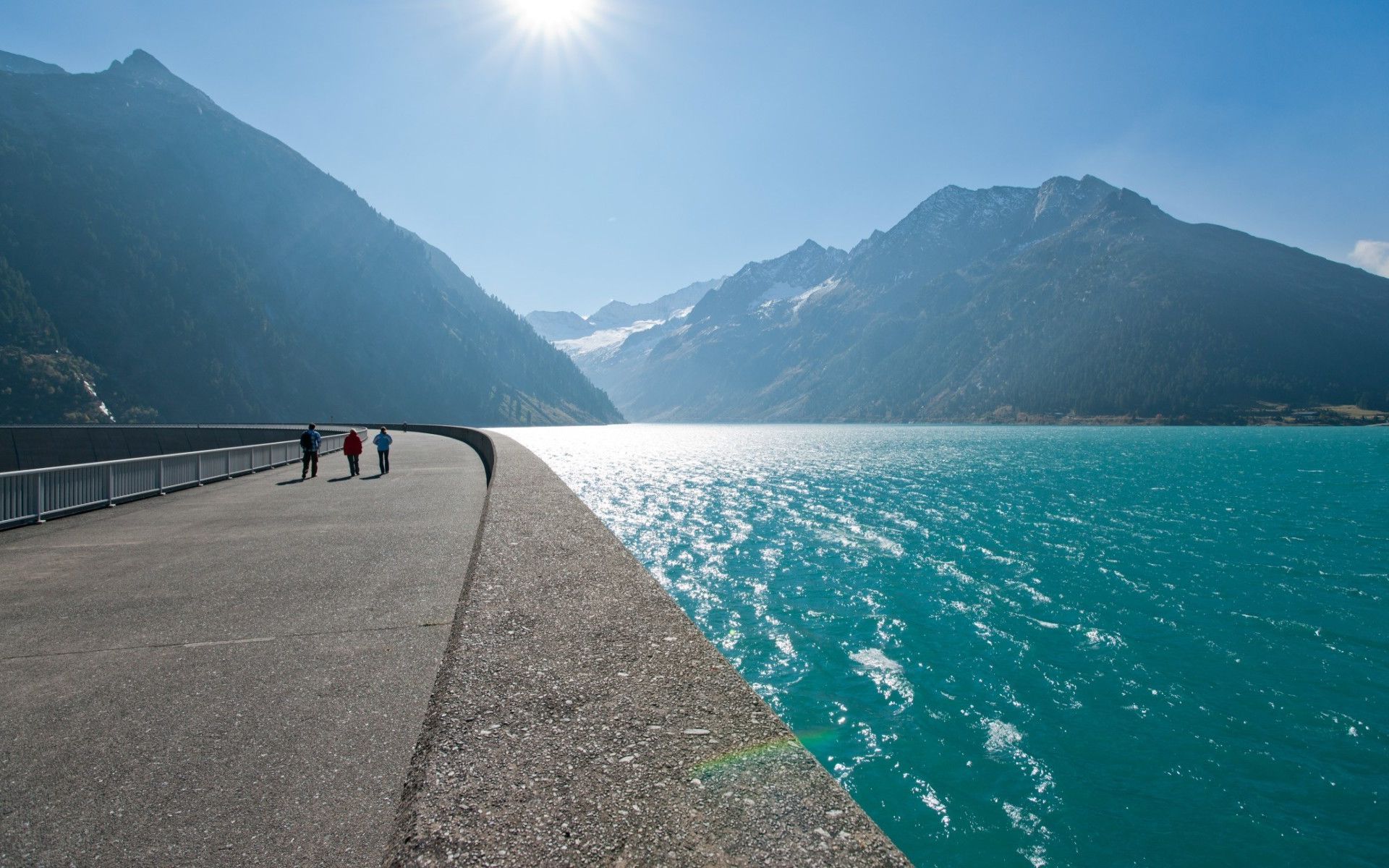 Staumauer des Schlegeisspeichers im Zillertal, Tirol. Rechts der türkisfarbene See, links hohe Berge. Menschen spazieren in der Sonne auf dem breiten Weg. Im Hintergrund schneebedeckte Alpen unter blauem Himmel.